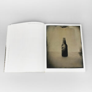 Open photo book showing a sepia-toned portrait of a single upright bottle on the right page; left page blank.