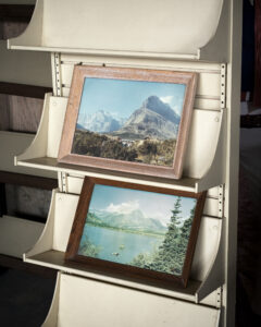 wo framed landscape paintings leaning on curved white display shelves, one above the other.