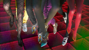 People's legs in tights, pants and fancy shoes above an abstract red board