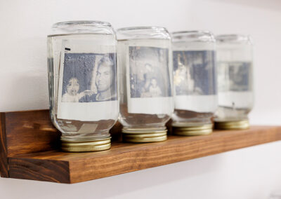 A shelf of four mason jars filled with liquid and polaroid photos.