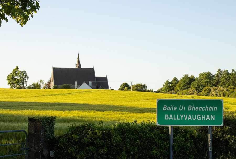 A photo of a landscape with a church and a road sign that says "Baile Uí Bheacháin Ballyvaughan."