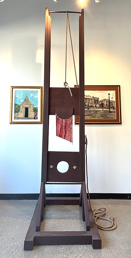 A large guillotine sculpture in front of two paintings.