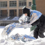 A person in a black snow suit dances with a reflective cloth in the snow.