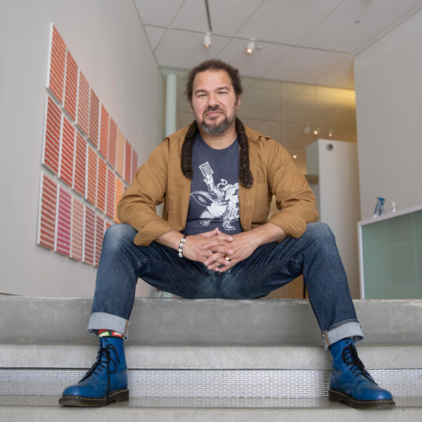 Man sits on staircase wearing with hands folded wearing brown jacket and blue jeans and sneakers