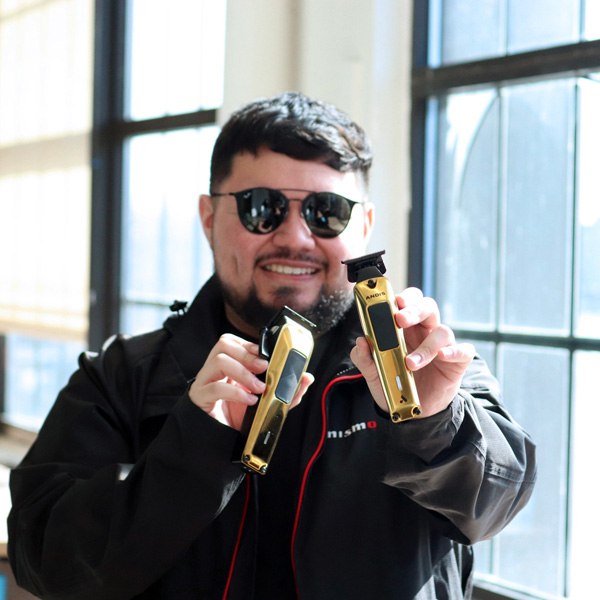 Young man wearing sunglasses holds two gold hair clippers and smiles.