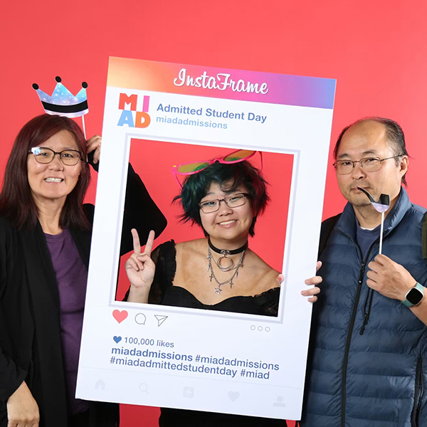 A student smiles and holds a large social media cutout frame while two adults stand holding props to the side.