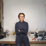 A man wearing a navy denim jumpsuit stands against a workbench full of tools and a sculpture