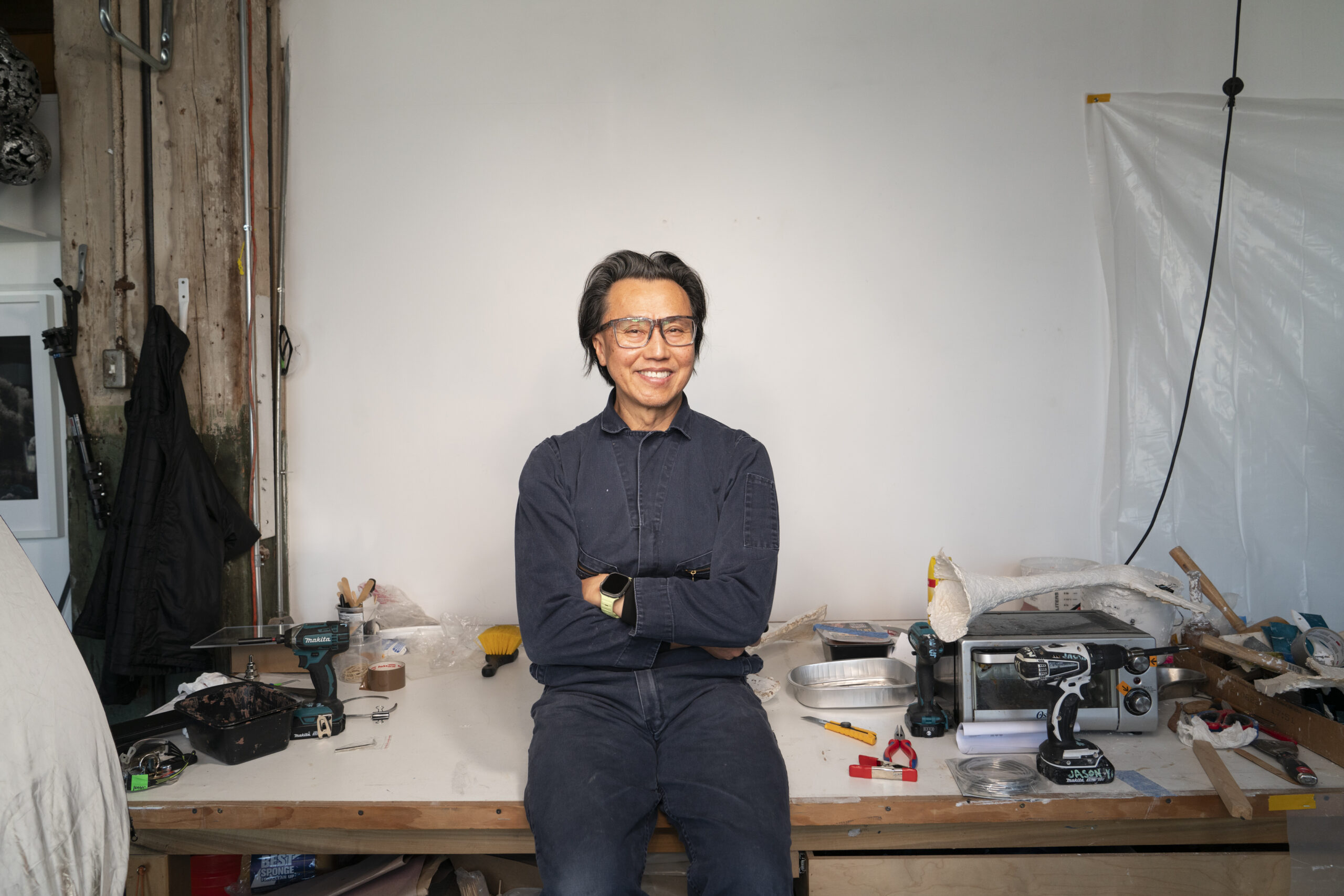 A man wearing a navy denim jumpsuit stands against a workbench full of tools and a sculpture