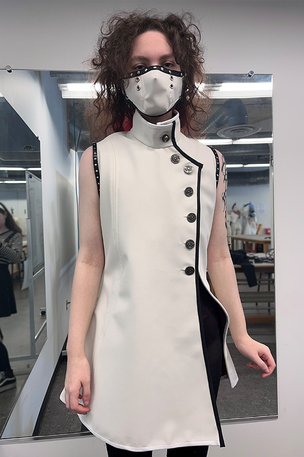A woman dressed in a sleeveless white asymmetrical jacket with black trim and matching face mask poses for a photo