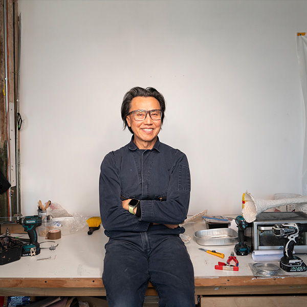 A man wearing a navy denim jumpsuit stands against a workbench full of tools and a sculpture