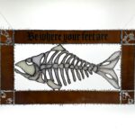 Stained-glass artwork of a fish skeleton framed by copper-toned panels with the phrase “Be where your feet are” across the top.