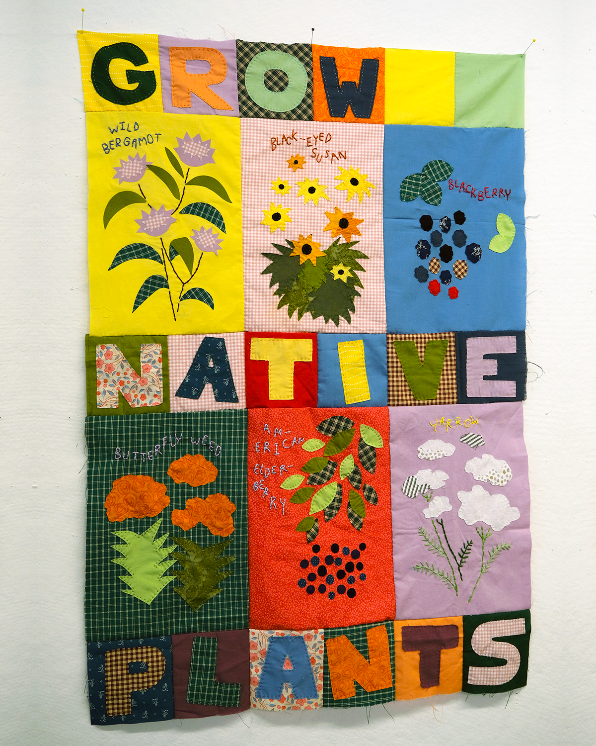Colorful quilt-style textile artwork featuring appliqué plants and large fabric letters spelling “GROW NATIVE PLANTS.” Each square panel includes stitched illustrations and handwritten labels for native plants including wild bergamot, black-eyed Susan, blackberry, butterfly weed, American elderberry and yarrow.