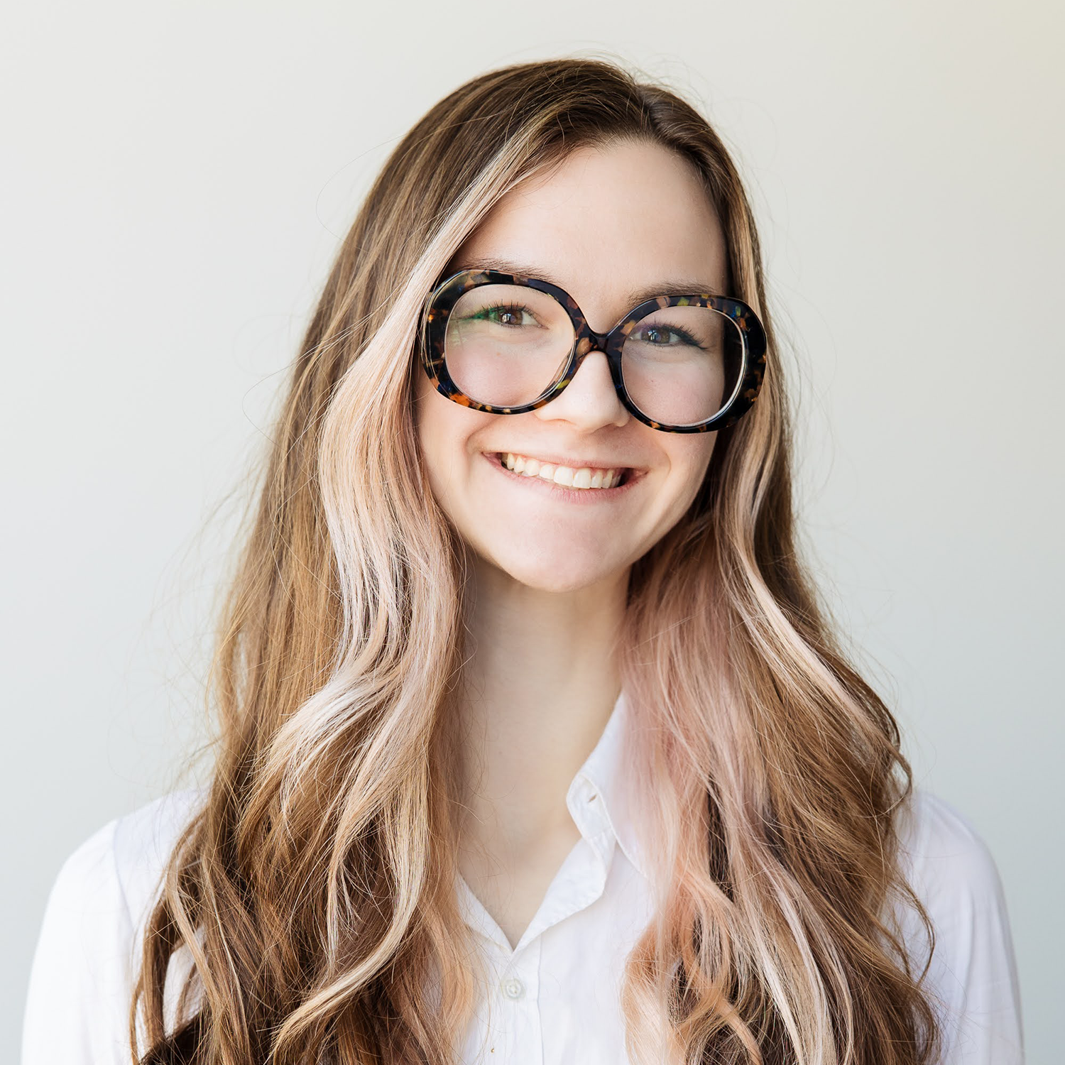 Kaitlyn Powers smiling with long wavy light brown hair and round tortoiseshell glasses wearing a white collared shirt against a light gray background.