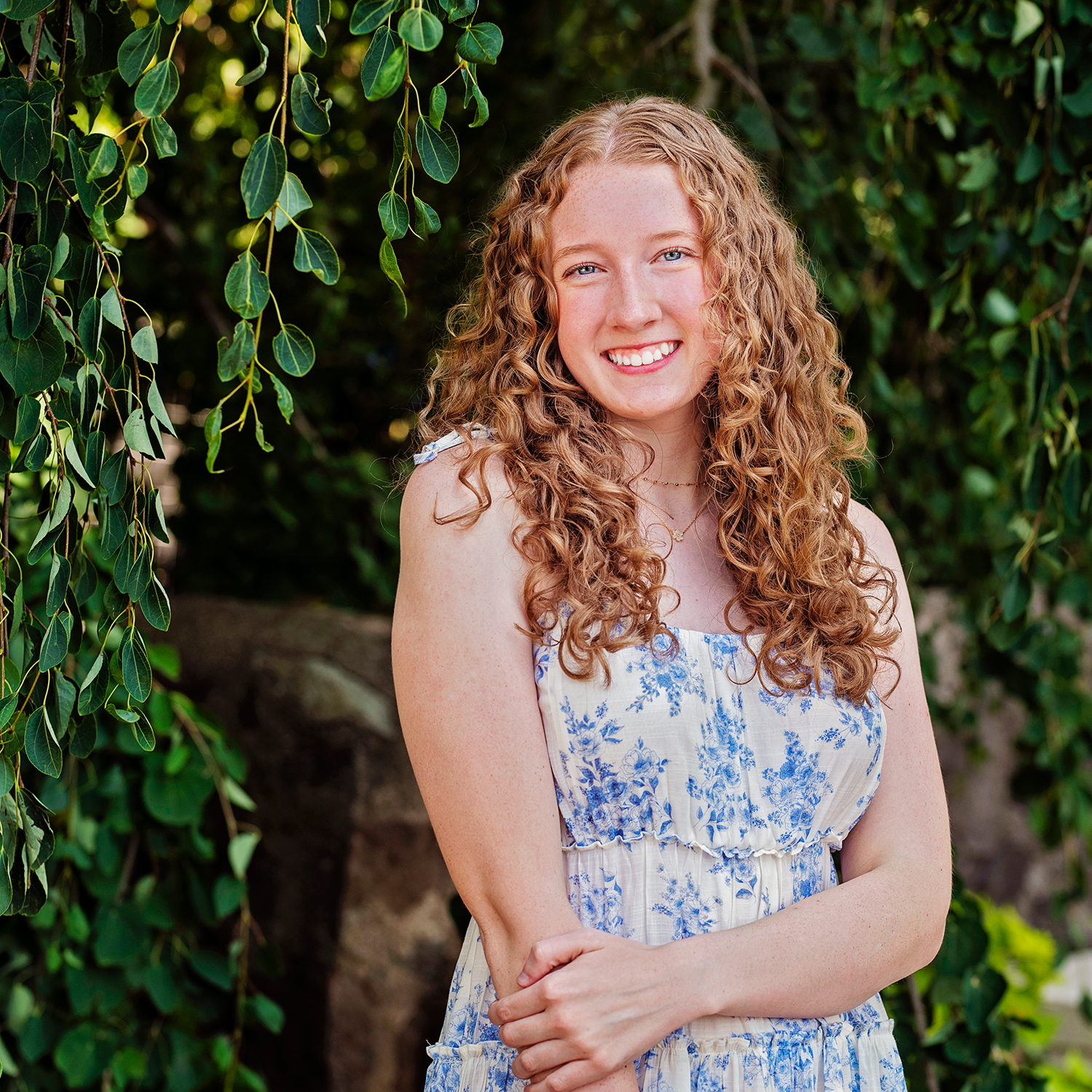 Sophia Simonson smiling with long curly red hair wearing a white and blue floral dress, standing outdoors in front of hanging green leaves and foliage.