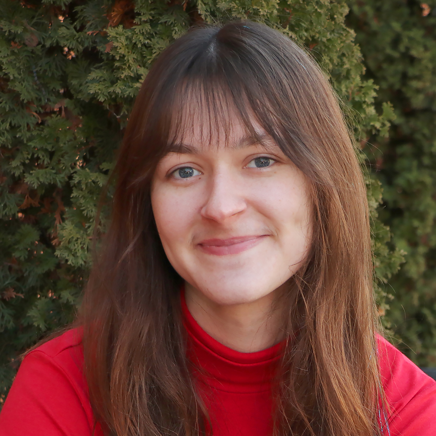 Portrait Natalie Spettel smiling with long brown hair and bangs, wearing a red turtleneck sweater, seated outdoors in front of evergreen foliage.