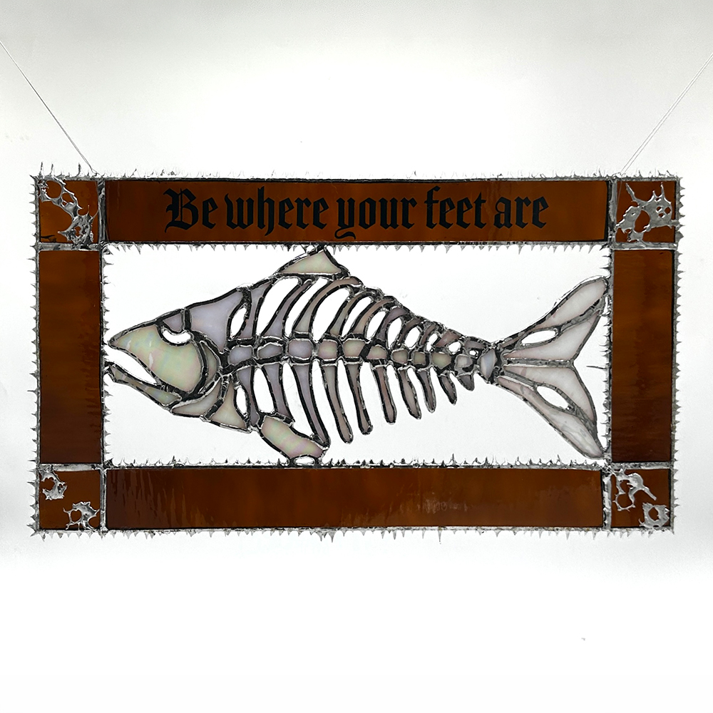 Stained-glass artwork of a fish skeleton framed by copper-toned panels with the phrase “Be where your feet are” across the top.