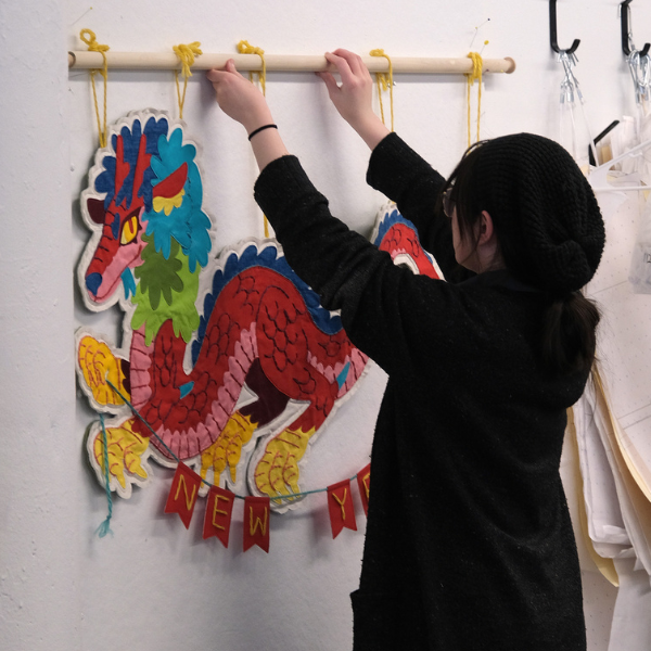 Person hanging a colorful textile dragon wall piece with a ‘New Year’ banner in a studio space.