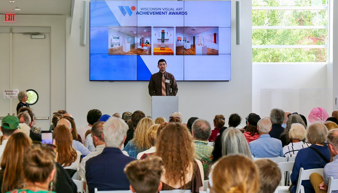 Chad Alexander Matha stands at a podium on a stage beneath a large screen showing the Wisconsin Visual Art Achievement Awards slide while a seated audience listens in a bright room.