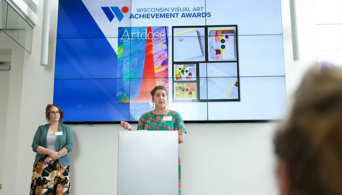 Rachel Hausmann Schall speaks at a podium in front of a large screen showing the Wisconsin Visual Art Achievement Awards logo and artwork while another person stands to the side.