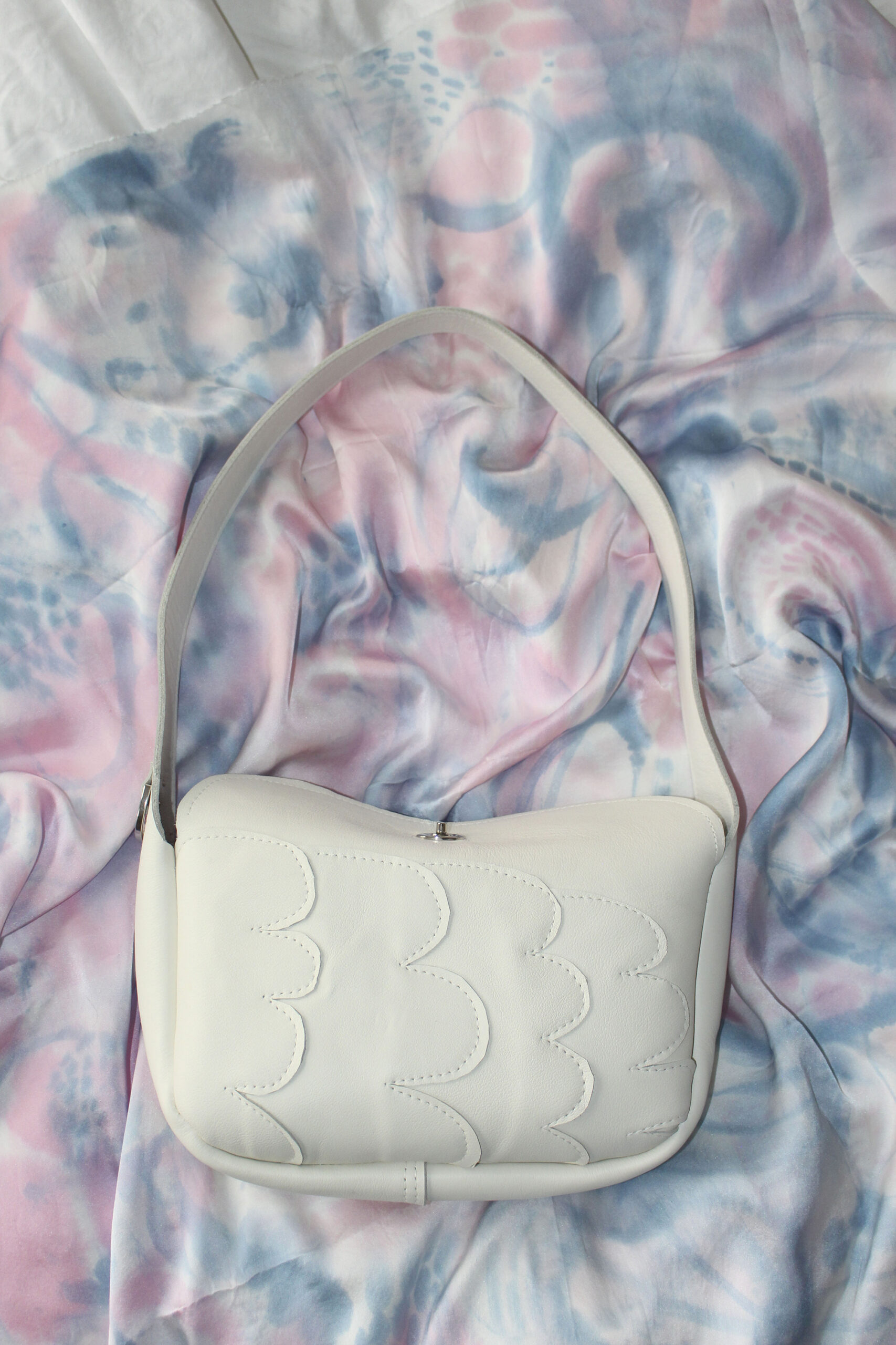 White leather shoulder bag with scalloped front detailing placed on a pastel pink and blue patterned fabric.