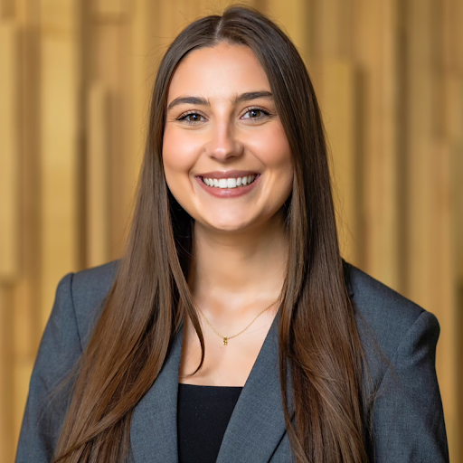 Head-and-shoulders professional portrait of Maya Koehn wearing a dark blazer and necklace, facing the camera against a warm-toned, softly blurred background with vertical architectural elements.