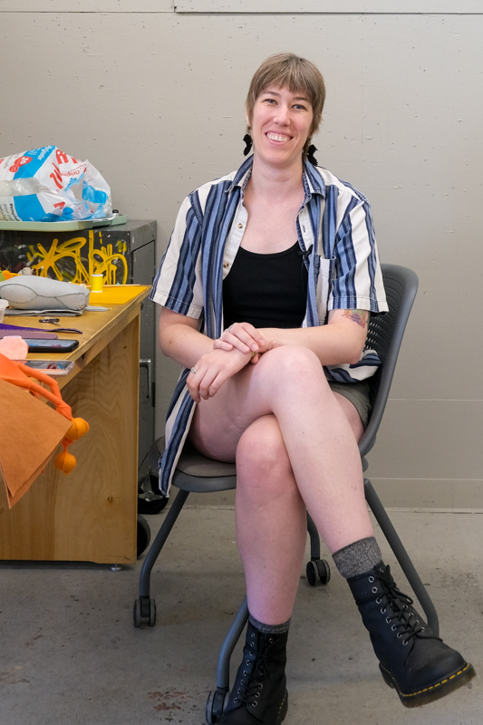 Gail Kowsky Person seated at a worktable surrounded by fabric, sewing materials, and handmade textile objects in an artist studio.