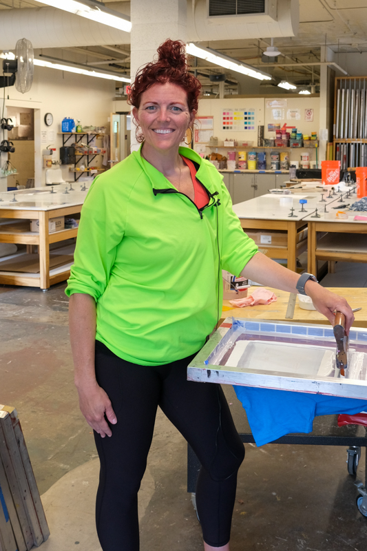 Kelly Wendel standing at a screen-printing station in a spacious printmaking studio.