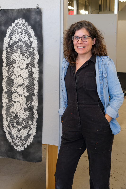 Alicia Zapata standing beside a display of textile artworks featuring lace-inspired floral patterns in an open studio workspace.
