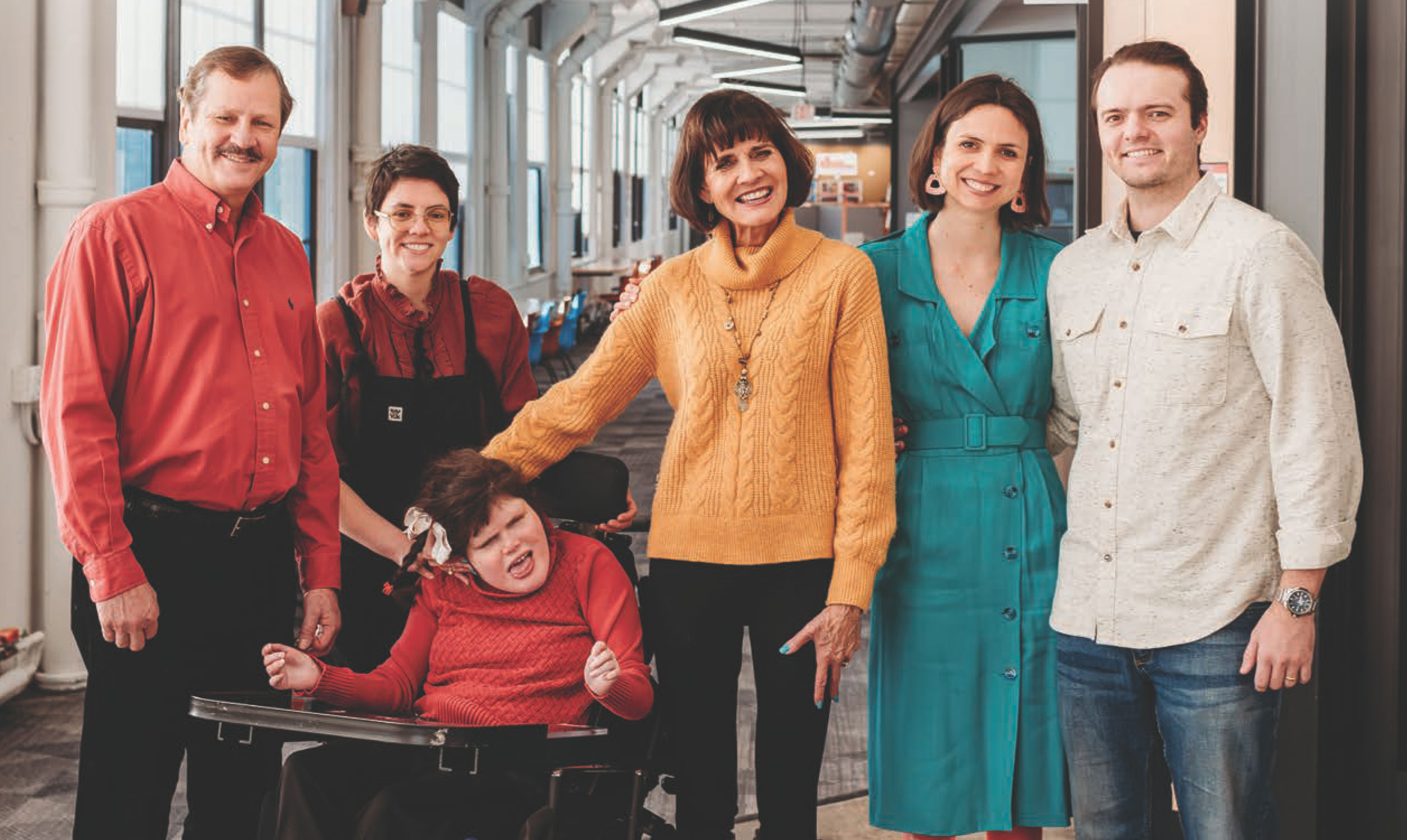 Group of five adults and one child using a wheelchair posed together in a bright indoor hallway with large windows.