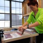 Kelly Wendel standing at a screen-printing station in a spacious printmaking studio.