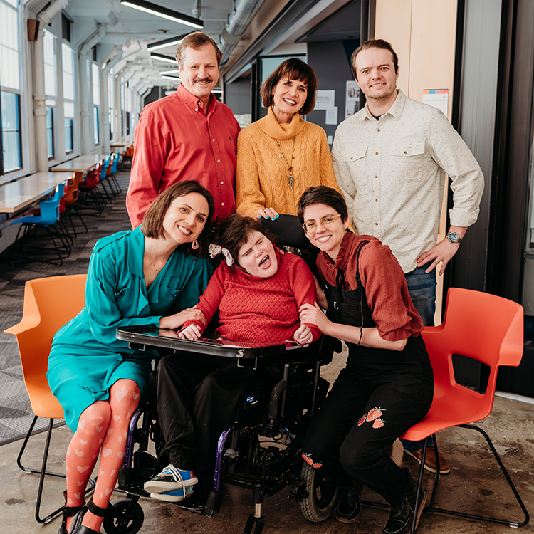 Six people gather around a wheelchair user in a bright campus common area with colorful chairs and large windows.