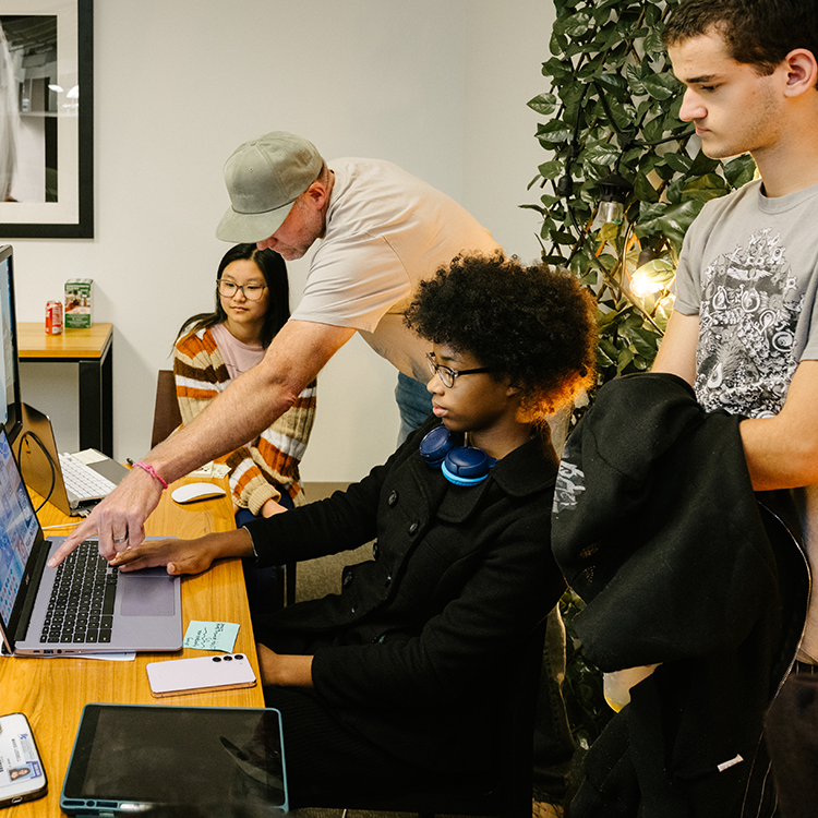 Students and an instructor review work on a laptop during a collaborative technology-focused learning session.