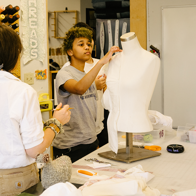 Students drape fabric on a dress form in a fashion design studio surrounded by sewing and patternmaking tools.