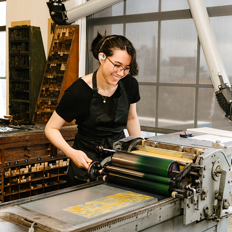 A student operates a printing press in a printmaking studio with type cabinets and workshop equipment nearby.