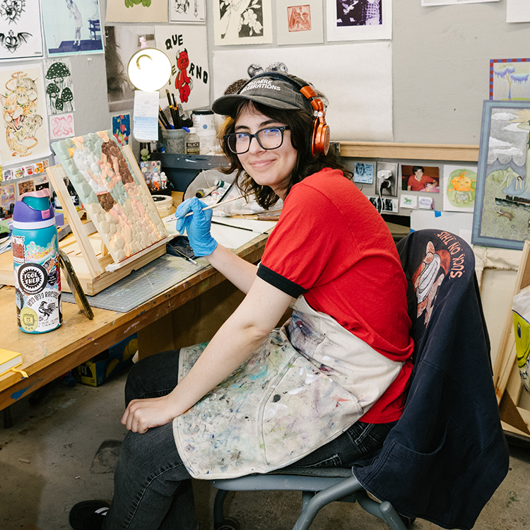 Student painting at a workbench surrounded by artwork, tools, and creative materials in a studio.