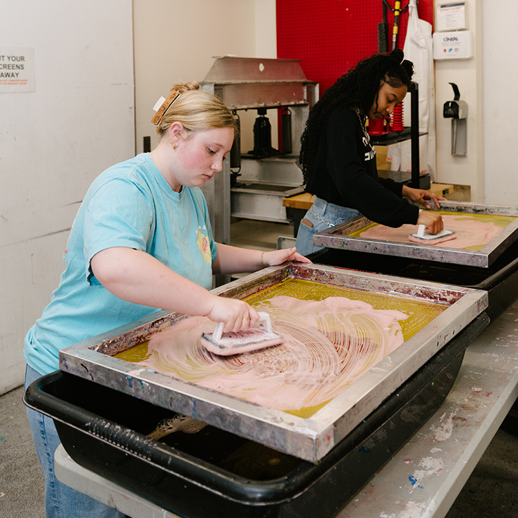 Students clean and prepare screen-printing frames at workstations in a printmaking studio.