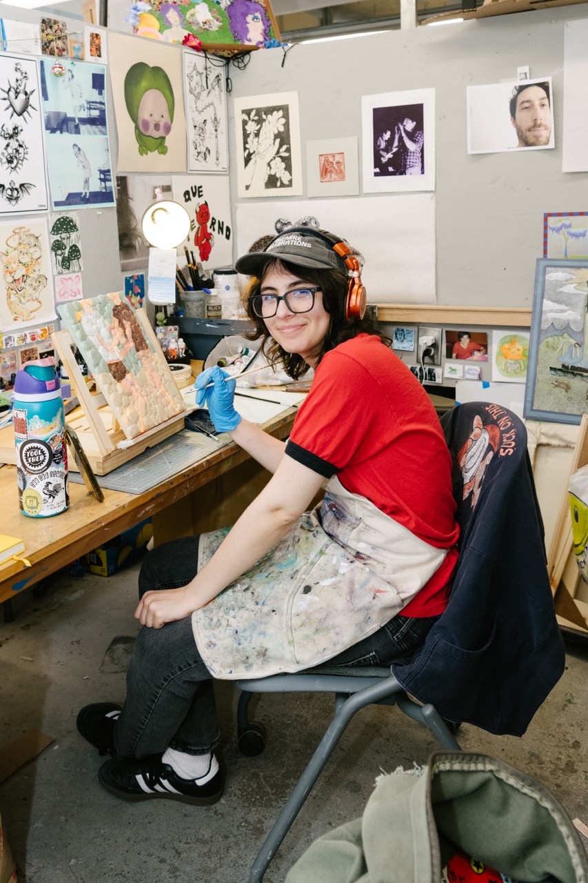 Student painting at a workbench surrounded by artwork, tools, and creative materials in a studio.