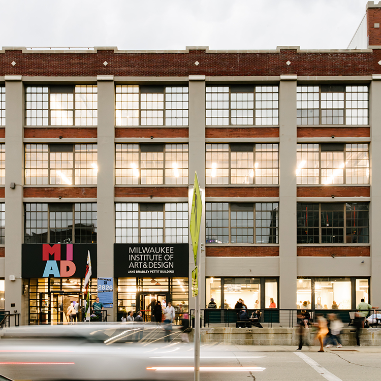 Exterior view of the Milwaukee Institute of Art & Design building along a city street, with pedestrians, large industrial windows, and traffic passing in front.