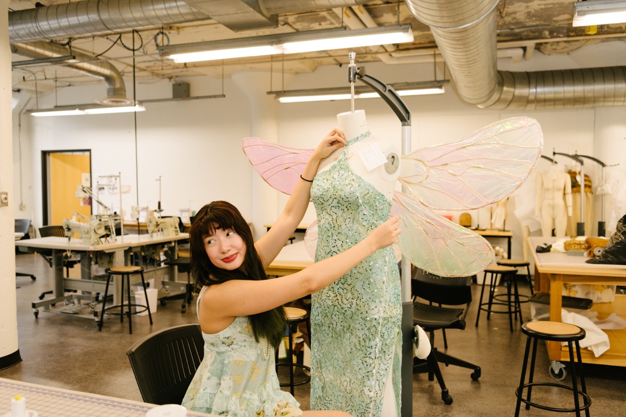 Student adjusts a sequined dress on a dress form with decorative translucent wings attached in a fashion design studio.