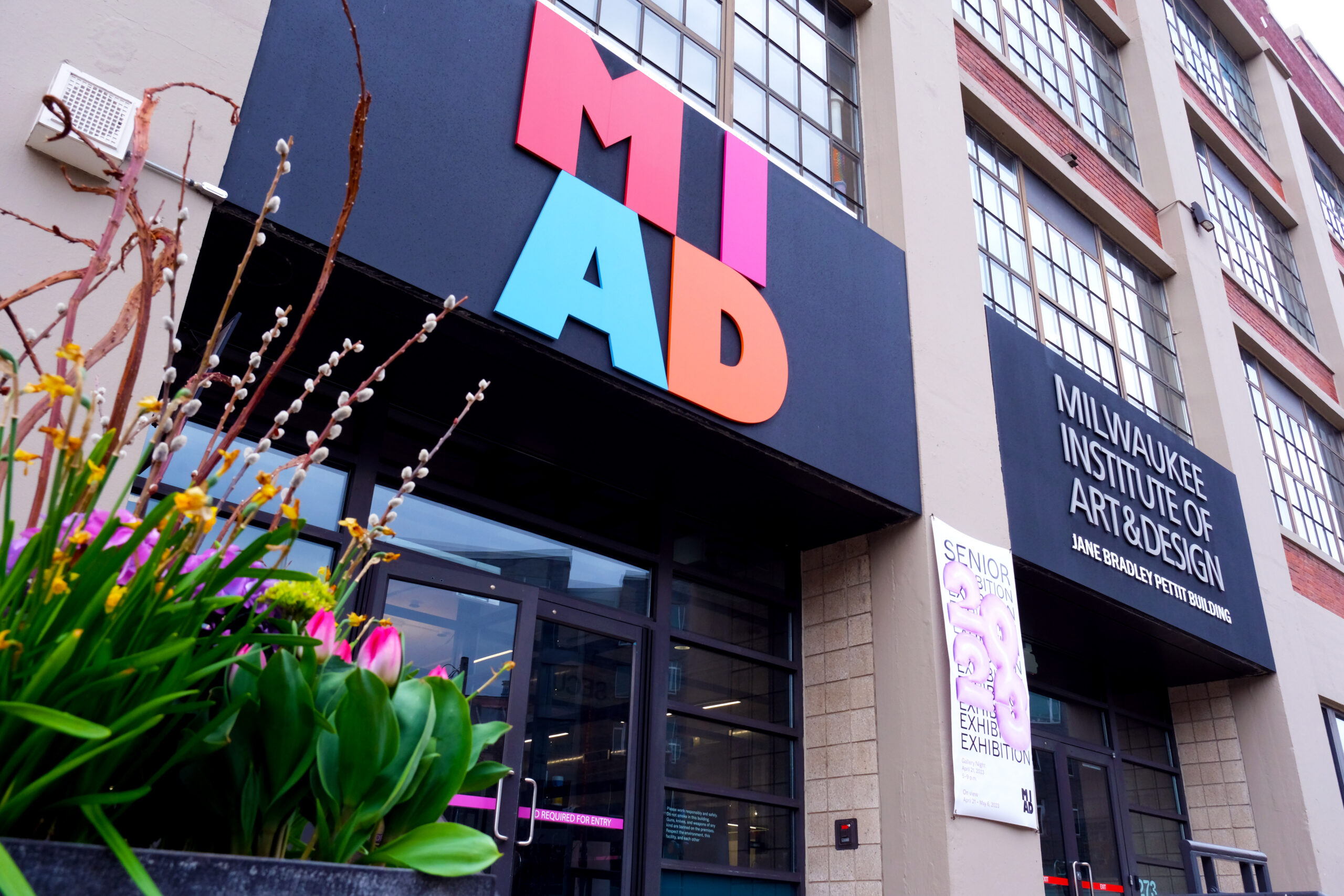 Entrance to the Milwaukee Institute of Art & Design building featuring a large colorful MIAD sign, tall industrial-style windows, and planters with flowers in the foreground.