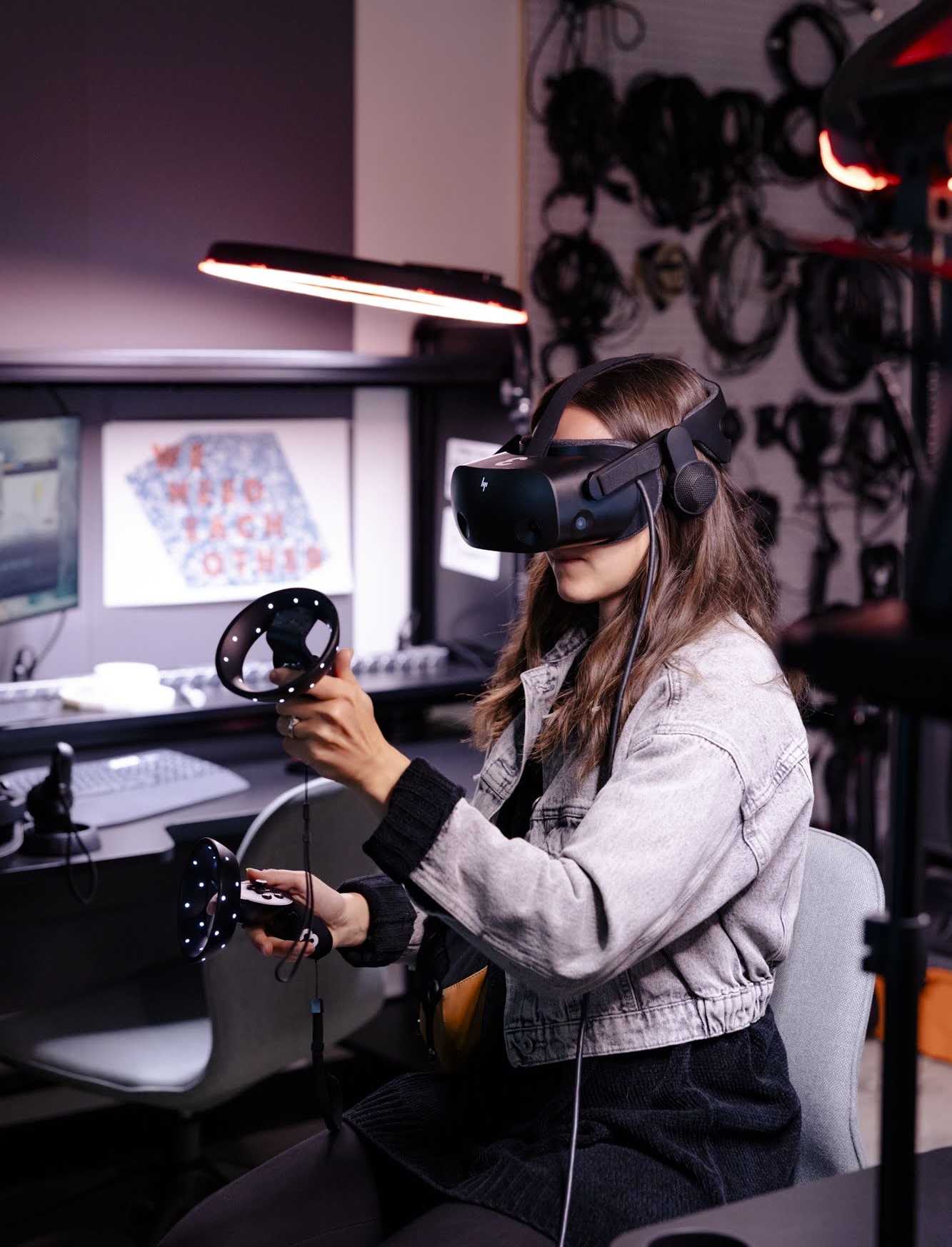 Student using a virtual reality headset and handheld controllers in a technology lab equipped with computers and digital media equipment.
