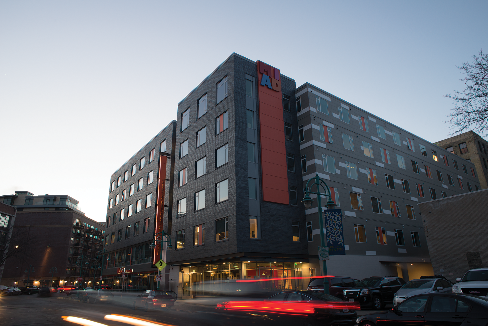 Exterior view of a multi-story MIAD student residence building at dusk, with illuminated windows and vehicle light trails along the street.