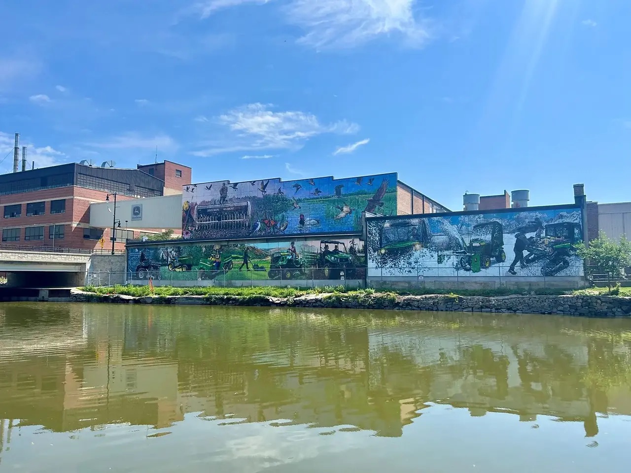 Large mural on industrial buildings along a riverfront depicting wildlife, wetlands, and John Deere equipment under a blue sky.