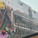 Mural panel featuring a yellow bird, wildflowers, and a historic John Deere Van Brunt agricultural seed drill.