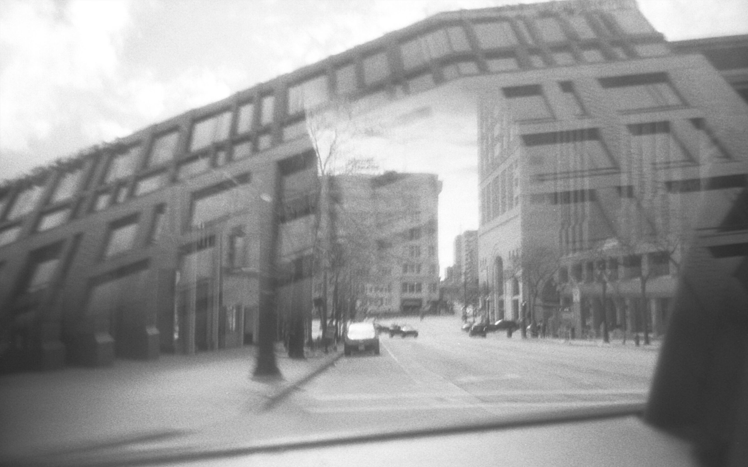 Black-and-white, soft-focus view of a city street lined with modern buildings, photographed through a window or reflective surface.