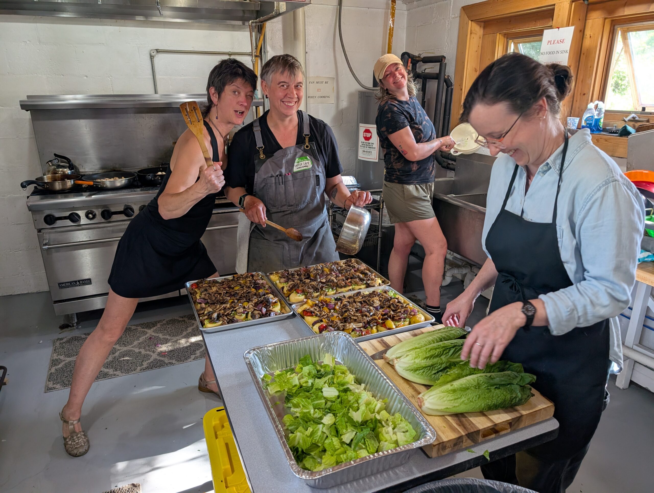 Four people preparing food together in a commercial kitchen, with trays of vegetables and lettuce on a worktable.