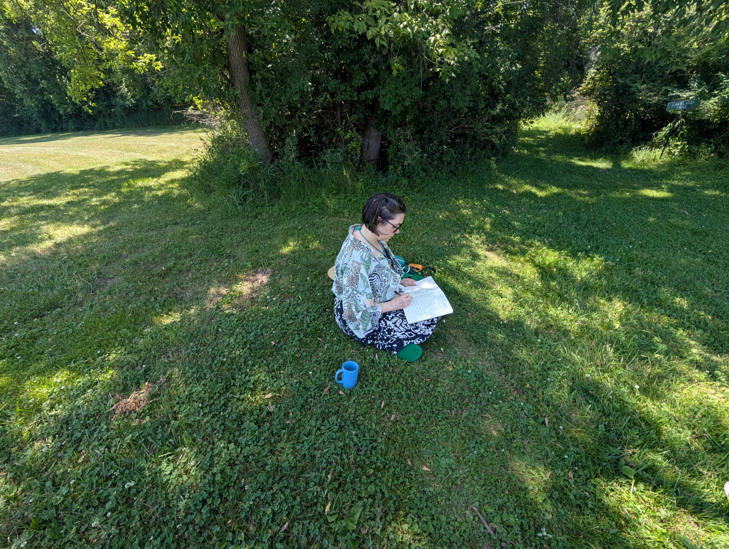 Person seated on grass under trees writing in a notebook during an outdoor retreat or study session.
