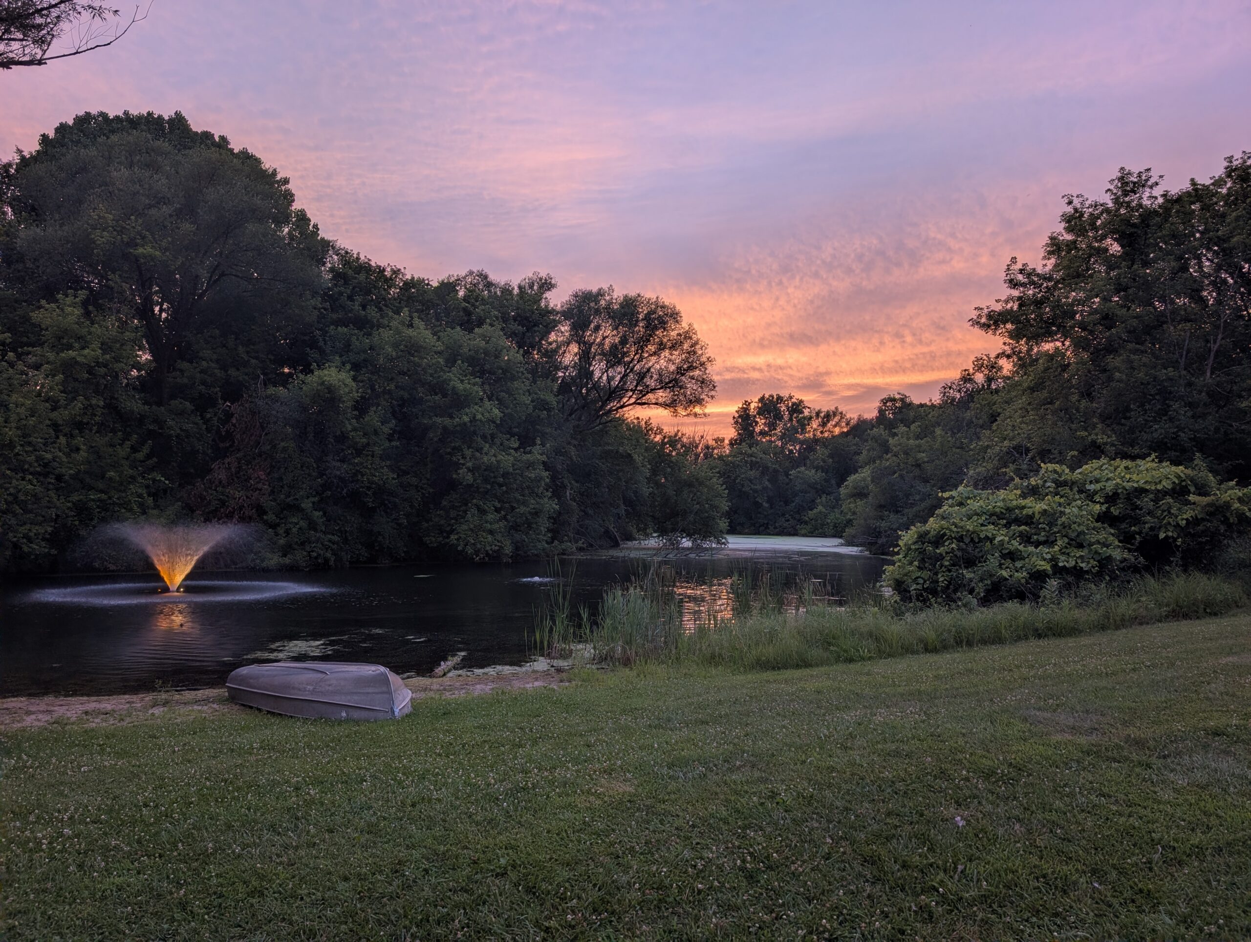 Pond surrounded by trees at sunset, with a small illuminated fountain and a rowboat resting on the grassy shore.