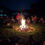 Group of people gathered around a campfire at night while music is played on acoustic instruments.