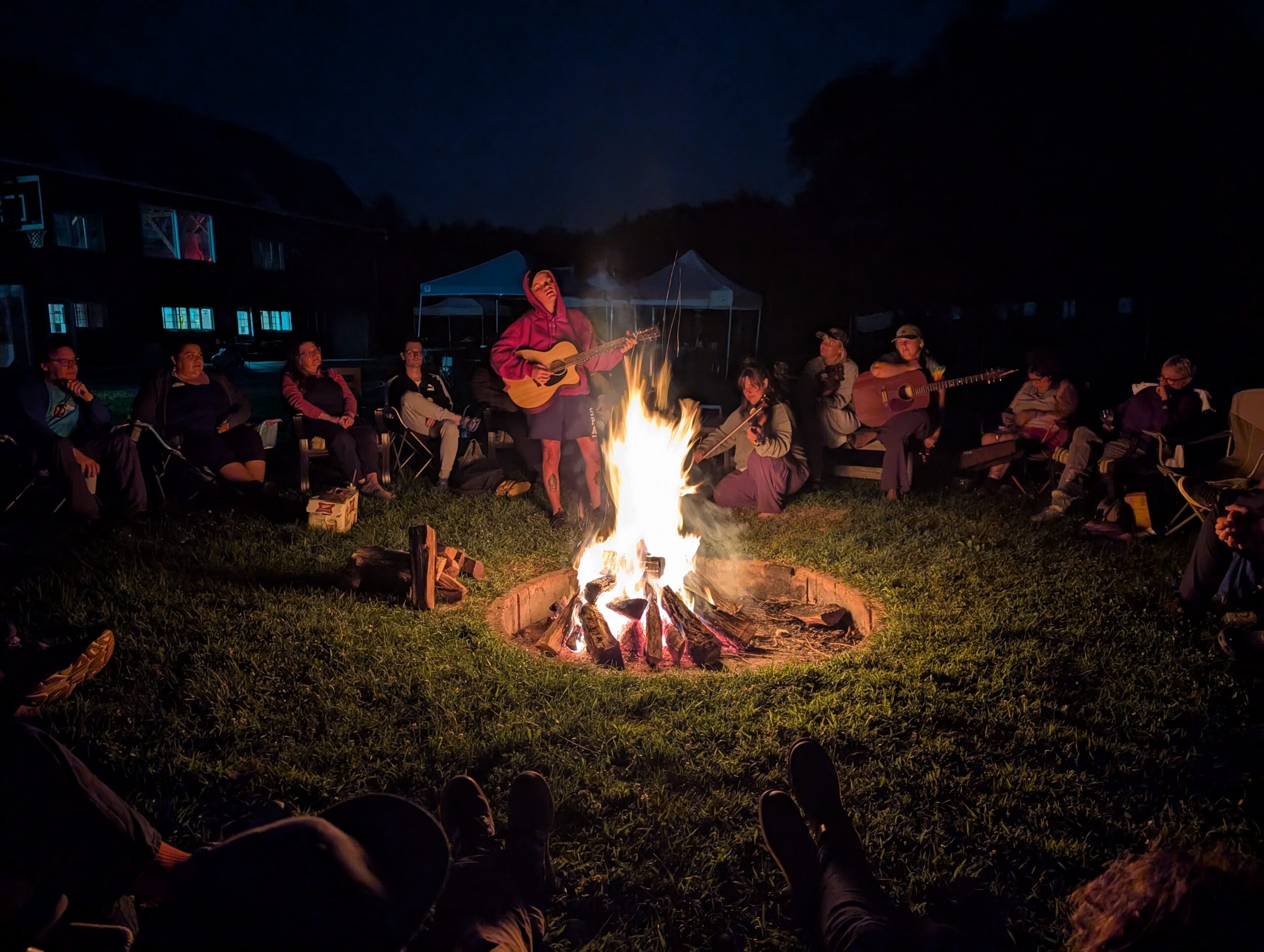 Group of people gathered around a campfire at night while music is played on acoustic instruments.
